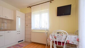 Una cocina pequeña con armarios blancos, un radiador, una ventana con cortinas blancas, una mesa con mantel floral y un televisor montado en la pared.