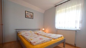 Una cama individual con colchón amarillo y ropa de cama estampada en una habitación con paredes de tonos claros y una ventana con cortinas traslúcidas.