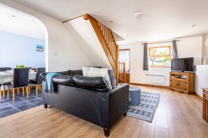 A modern living room with a black leather sofa, a wooden staircase, a dining area with a checkered tablecloth, and a television on a wooden cabinet. - Y Stabl: Pet-friendly cottage with stunning views (Ty Cros)