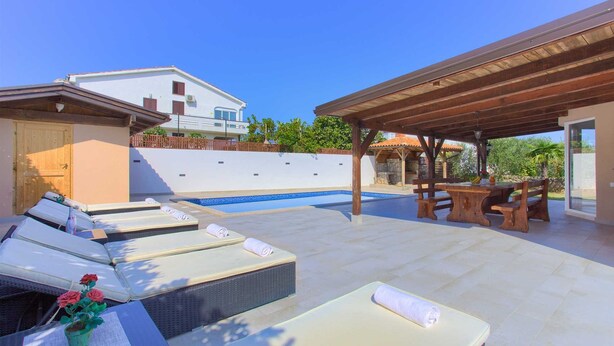Zona junto a la piscina con sillas de descanso, un área de estar cubierta y vista a un edificio al fondo.