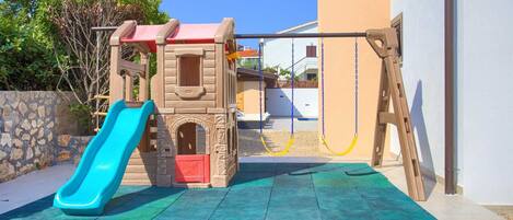 Un espace de jeux pour enfants avec un toboggan et des balançoires, situé devant un bâtiment doté d’un mur en pierre et sous un ciel dégagé.