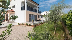 Una casa moderna de dos pisos con balcón, camino de entrada de grava y vegetación circundante.