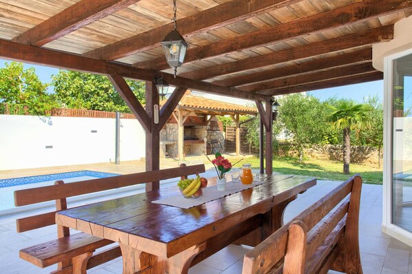 Un espace de restauration en plein air avec une table et des bancs en bois, un luminaire suspendu, et une vue sur une piscine et un jardin.