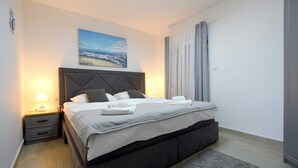 Una cama bien hecha con almohadas y una manta, una mesita de noche con una lámpara, un cuadro en la pared y una ventana con cortinas.