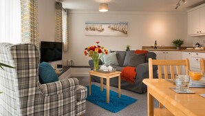 A living room with a sofa, a coffee table with flowers, a TV, and a kitchen area with cabinets and a sink. - Ty Arthur: Ground-Floor Apartment for Two in Aberffraw | Managed by Anglesey Holiday Lettings (Ty Croes)
