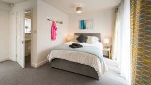 A hotel room with a bed, bedside lamps, a headboard, a wall-mounted artwork, and a door leading to a bathroom. - Ty Arthur: Ground-Floor Apartment for Two in Aberffraw | Managed by Anglesey Holiday Lettings (Ty Croes)