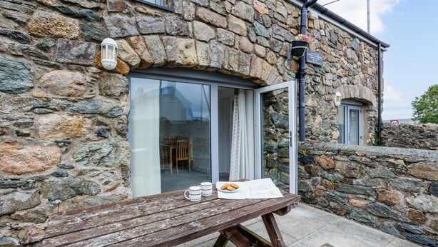 A stone building with a wooden bench, a table with a newspaper and two mugs, and a glass door leading inside. - Ty Arthur: Ground-Floor Apartment for Two in Aberffraw | Managed by Anglesey Holiday Lettings (Ty Croes)
