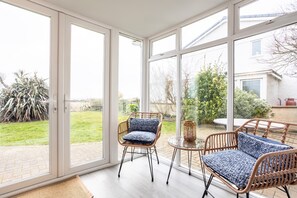 A conservatory with wicker furniture and a glass table, overlooking a garden. - Lakeside : Stylish 4‑Bed Family Villa in Rhosneigr | Managed by Anglesey Holiday Lettings (Rhosneigr)