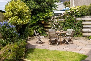 A patio with a wooden table and chairs, surrounded by lush greenery and flowering plants.