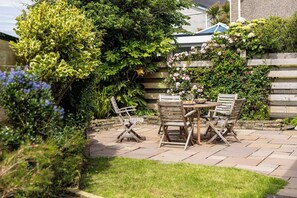 Eine Terrasse mit Holztisch und -stühlen, umgeben von üppigem Grün und blühenden Pflanzen.