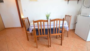 Uma área de refeições com uma mesa e cadeiras de madeira, uma pequena planta sobre a mesa, e uma cozinha com eletrodomésticos e uma prateleira acima.