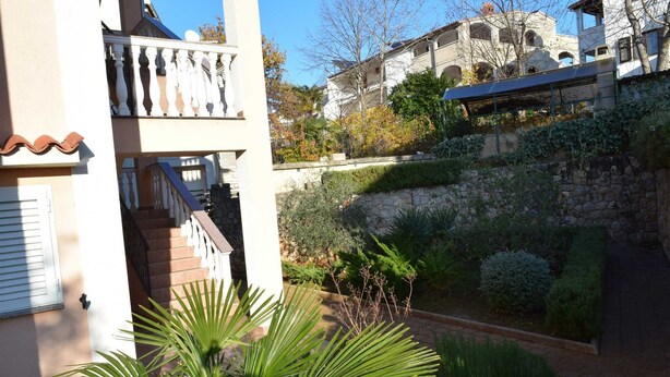 Área residencial com uma escadaria de pedra que leva a uma varanda, cercada por vegetação exuberante e um muro de pedra.