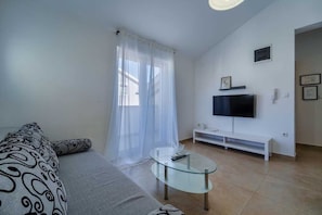 Un salon avec un canapé, une table basse en verre, un téléviseur posé sur un meuble et une fenêtre ornée de rideaux légers.