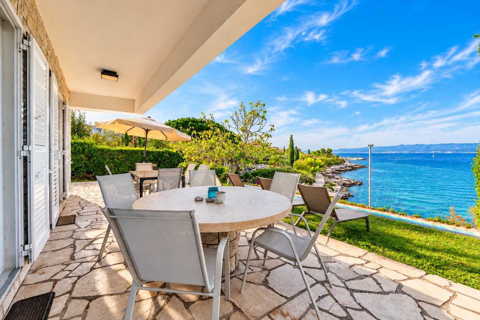 Eine Terrasse im Freien mit einem runden Tisch und Stühlen, mit Blick auf ein Gewässer und einen klaren Himmel.