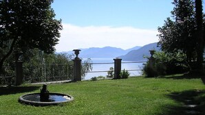 Un espace verdoyant agrémenté d’une fontaine, de piliers en pierre et offrant une vue sur un lac et des montagnes.