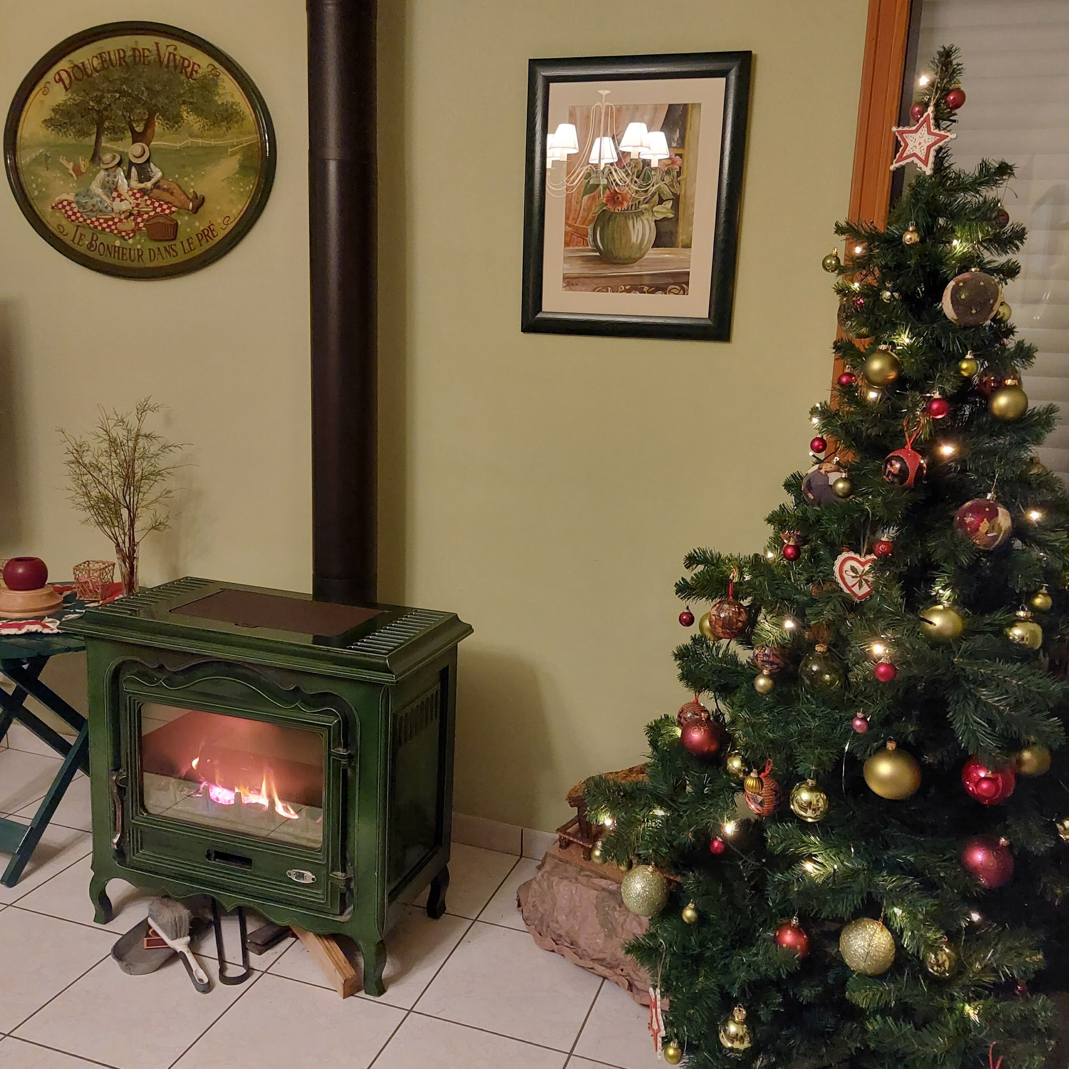 A cozy room with a green wood-burning stove, a Christmas tree decorated with ornaments, and a framed picture on the wall.