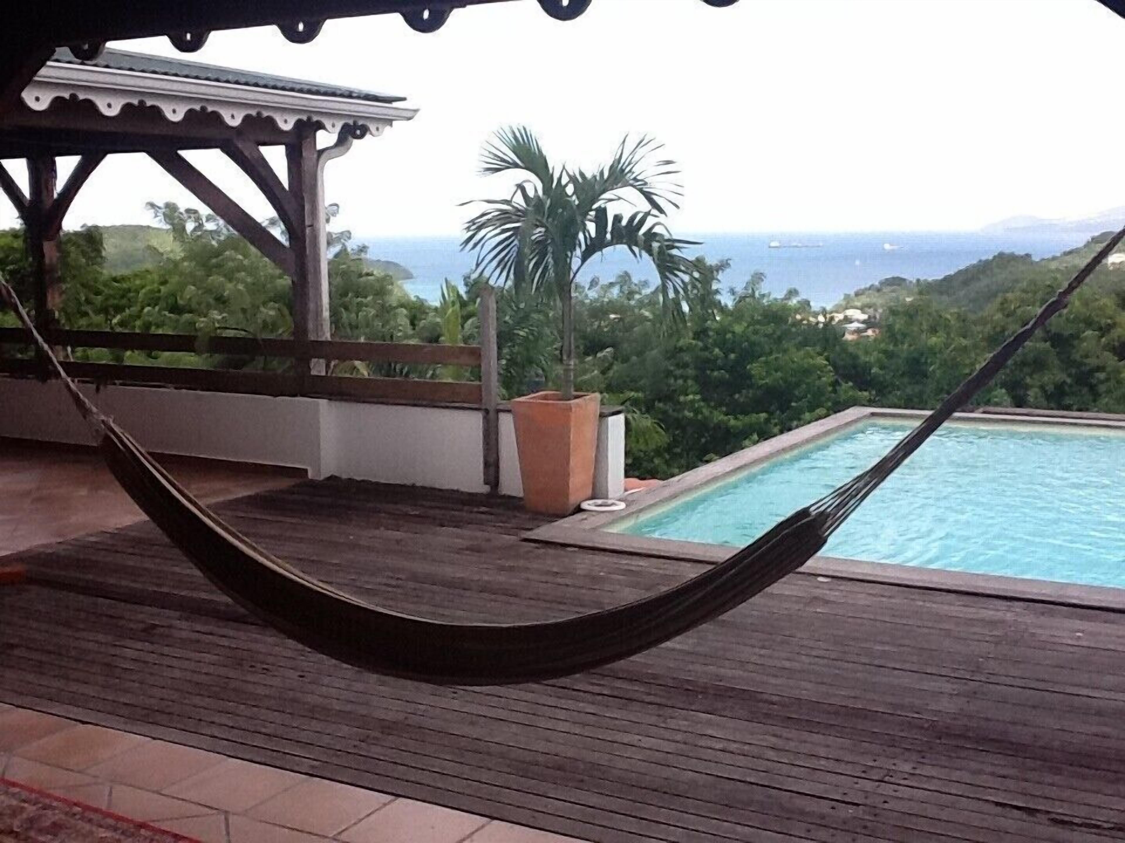 A hammock on a wooden deck overlooking a swimming pool with a view of the ocean and greenery.