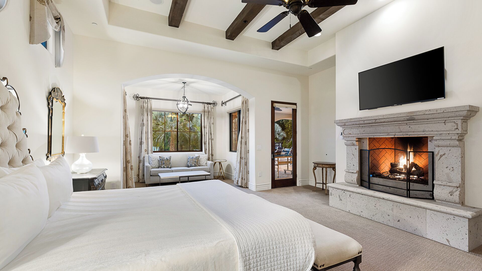 A spacious bedroom with a large bed, a fireplace, a television mounted above it, and a view of the outdoors through a window.
