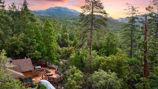 Una cabaña con terraza y zona de estar al aire libre, rodeada de un bosque denso y montañas.