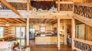 Interior espaçoso de cabana, com cozinha central, paredes de madeira e um couro de vaca pendurado.