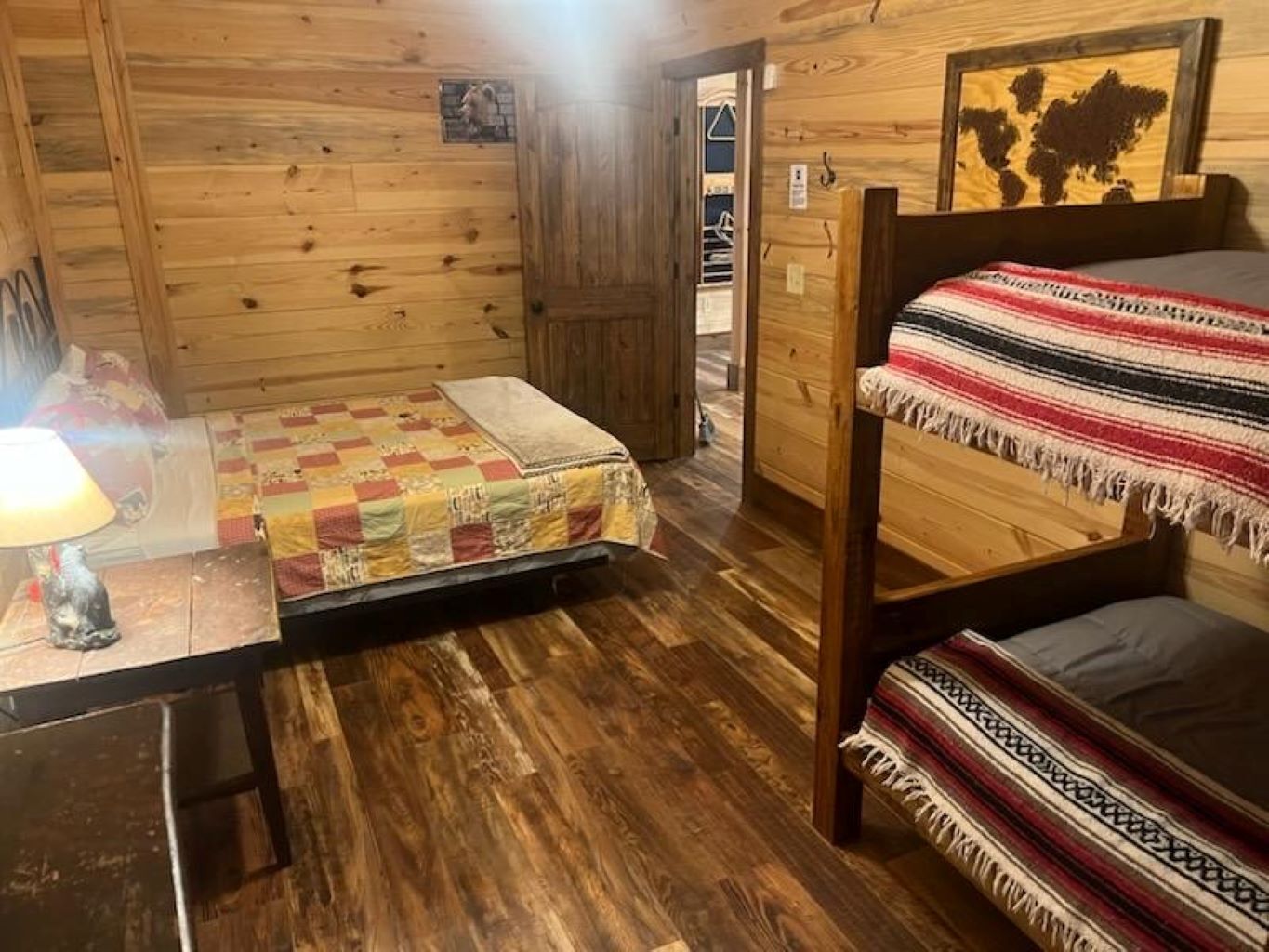 A cozy cabin room with a wooden floor, a bed with a colorful quilt, a small table with a lamp, and a wooden bunk bed.