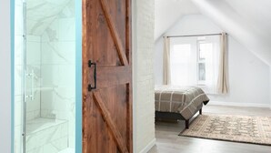 Un dormitorio con cama, puerta de madera, ducha de vidrio y ventana con cortinas.