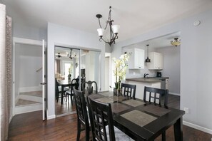 A dining area with a dark wooden table and chairs, a chandelier, and a kitchen in the background.