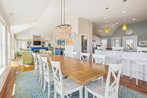 A spacious dining area with a wooden table and white chairs, a kitchen with white cabinets, and a living area with a blue sofa. - OCEANFRONT!  ELEVATOR!   HOTTUB!   5 Masters!   AMAZING OCEAN VIEWS! (Duck)