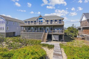 A two-story beachfront house with a balcony, a covered deck, and a sandy pathway leading to the entrance. - OCEANFRONT!  ELEVATOR!   HOTTUB!   5 Masters!   AMAZING OCEAN VIEWS! (Duck)