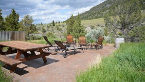 An outdoor patio with picnic tables and chairs, surrounded by grass and trees, overlooking a hilly landscape. - Horse Ranch on the Yellowstone River. Close to Gardiner and Yellowstone Park. (Emigrant)