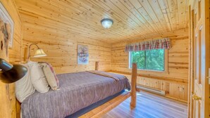 A wooden cabin bedroom with a bed, a nightstand, a lamp, and a window with a view of trees. - Horse Ranch on the Yellowstone River. Close to Gardiner and Yellowstone Park. (Emigrant)