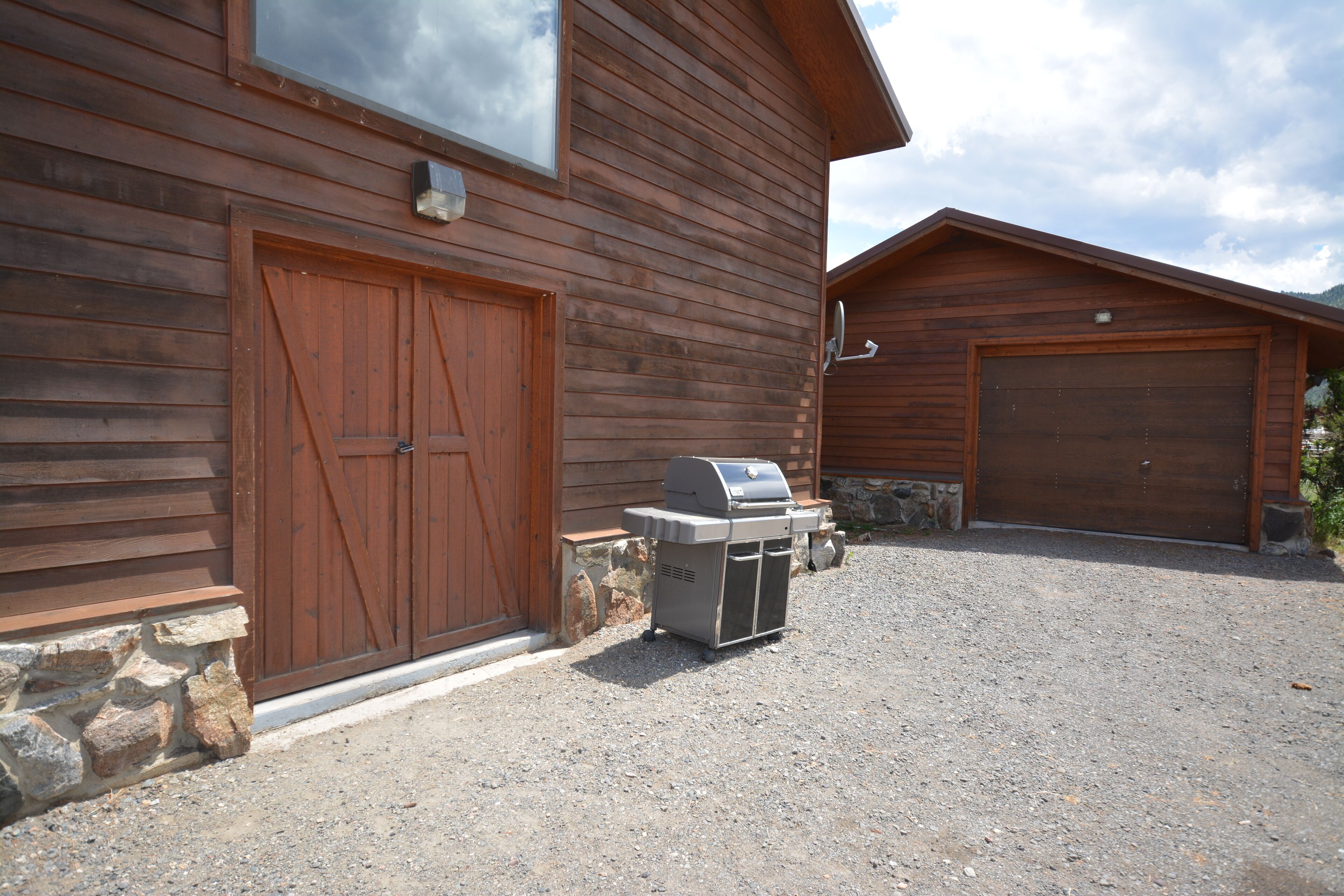 A wooden building with two garages, a stone foundation, and a barbecue grill in front.