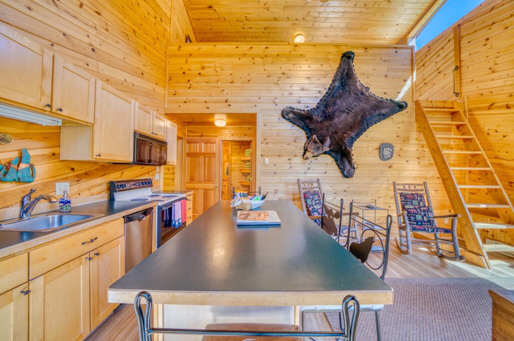 A wooden interior with a central dining table, kitchen cabinets, a mounted animal hide, and a ladder.