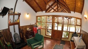 A cozy room with wooden floors, a green chair, a table with a bottle, and a large window offering a view of the outside. - Hostal de la Laguna (Ushuaia)