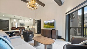 Uma sala de estar moderna com sofá, mesa de centro e área de jantar. Cozinha visível ao fundo, com uma janela grande.