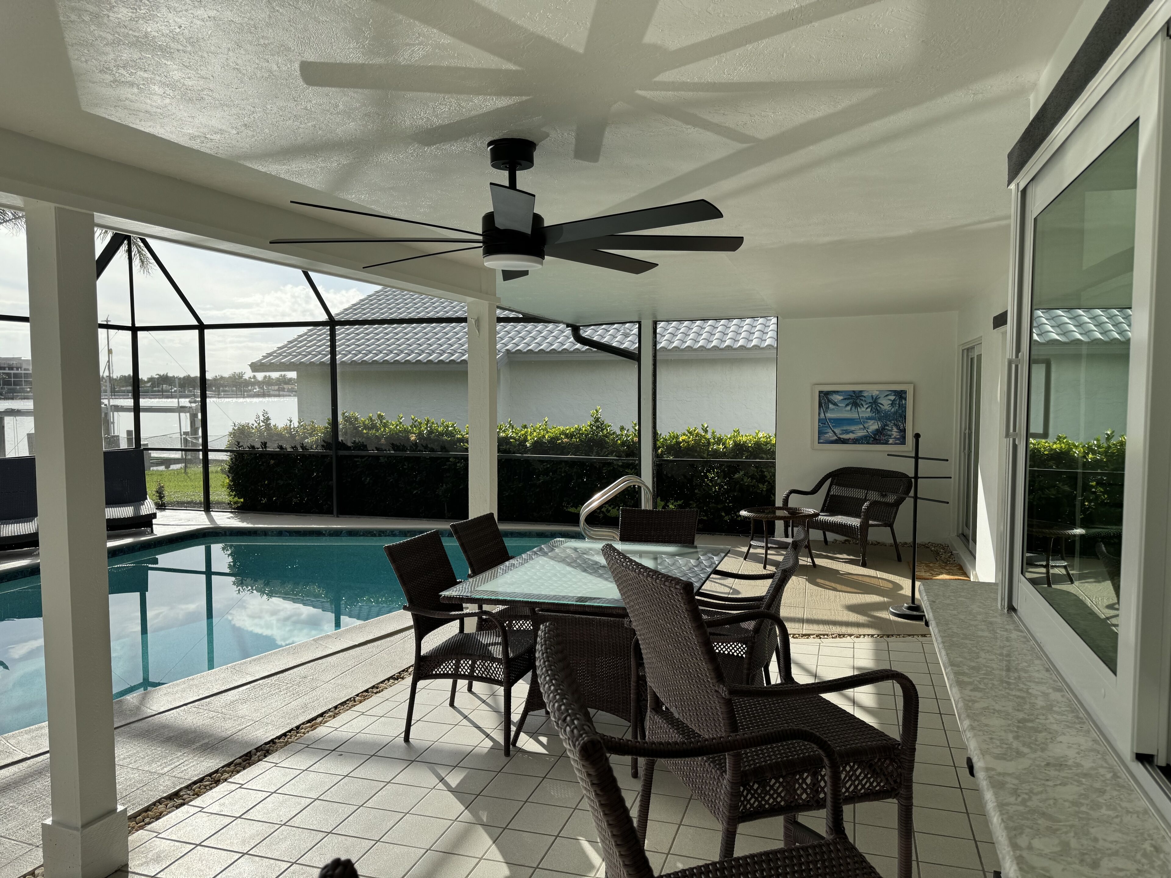 A poolside area with a glass table and wicker chairs, a ceiling fan, and a view of a building and greenery.