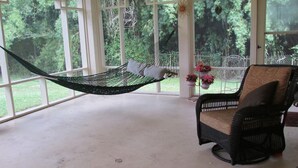 A hammock suspended between white-framed windows, a wicker chair with a cushion, and potted plants on a balcony with a view of greenery.