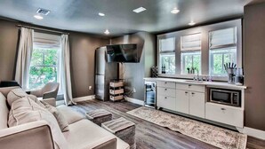 A modern living room with a sofa, ottomans, a flat-screen TV, a kitchen with a microwave, and a window with curtains.