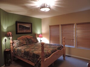 Quarto com cama de madeira, mesa de cabeceira, luminária, quadro na parede e janela com persianas.