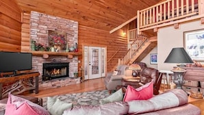 A log cabin interior with a stone fireplace, a sofa with cushions, a television, and a staircase. - Serenity Retreat (Formerly Leins Lodge) Luxury Home Stunning Mountain Views (Maggie Valley)