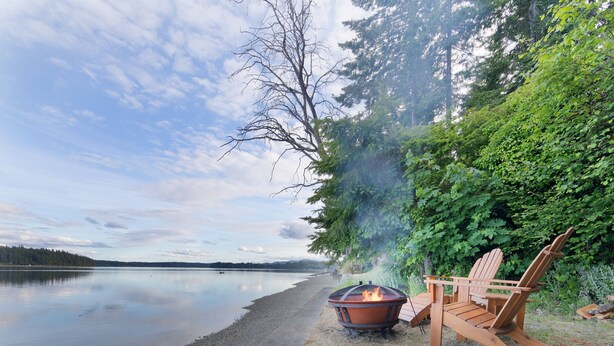 Un cadre idyllique au bord du lac, avec un foyer et des chaises en bois installés près de l’eau.