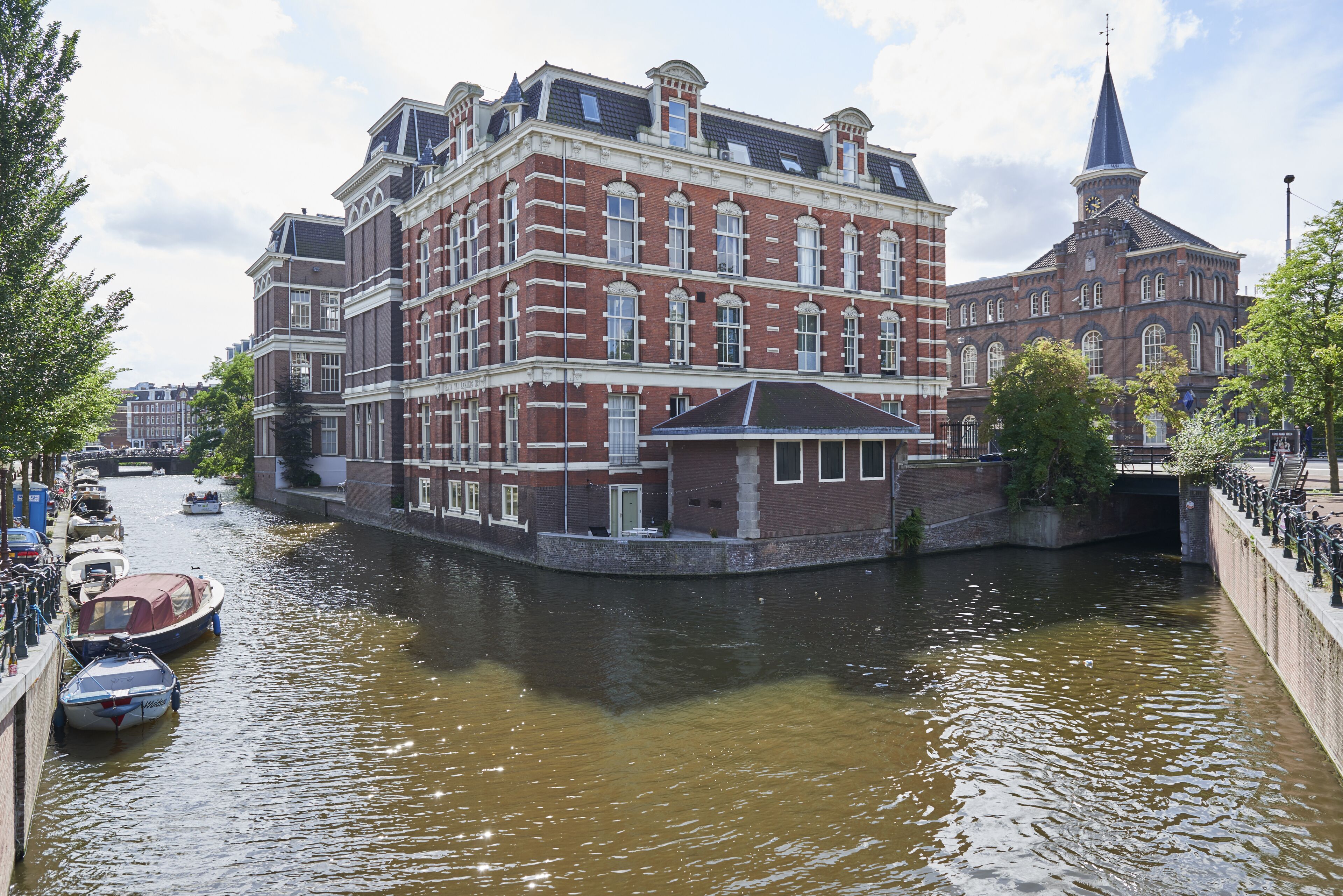 Un edificio junto a un canal con una destacada torre del reloj, un puente cubierto y una hilera de barcos amarrados a la orilla.