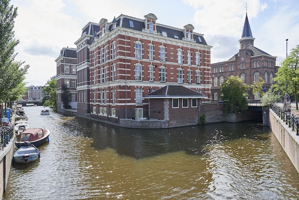 Un bâtiment situé au bord du canal, doté d’un imposant clocher, d’un pont couvert et d’une rangée de bateaux amarrés le long de la berge.