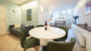 A modern living room with a round dining table, green chairs, a sofa with patterned wallpaper, and a kitchen area in the background.
