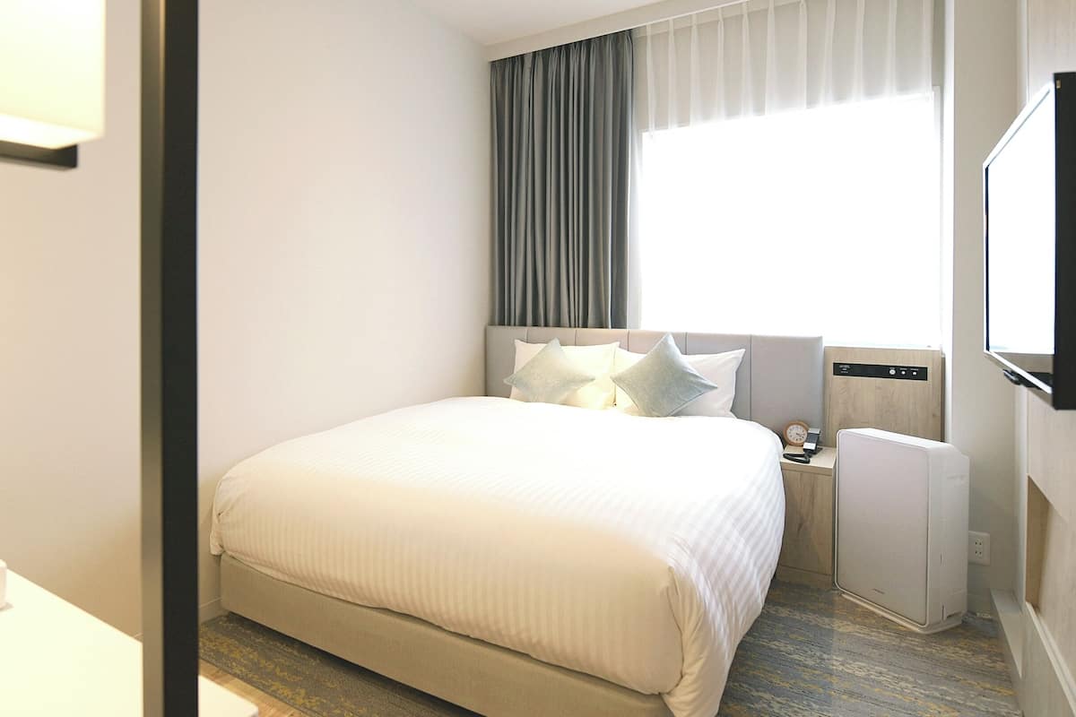 A hotel room with a large bed, a bedside table with a lamp, a TV, and a window with curtains.