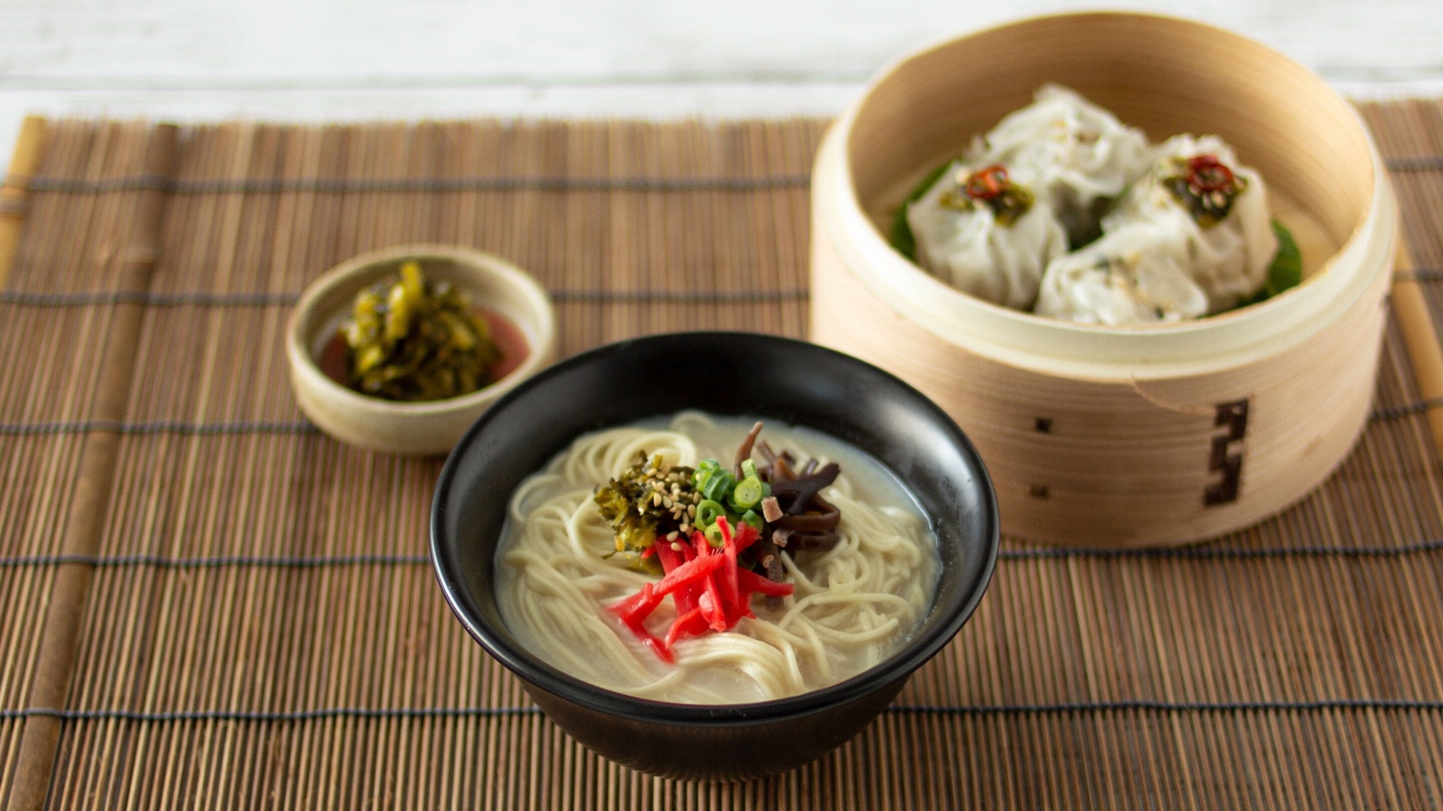 A bowl of ramen with toppings, a small bowl of pickled vegetables, and a bamboo steamer with dumplings.