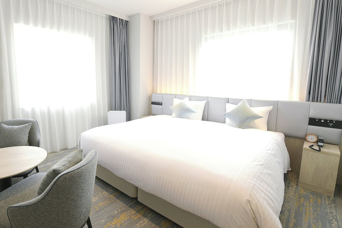 A hotel room with a large bed, a small table, two chairs, and a bedside table with a clock.