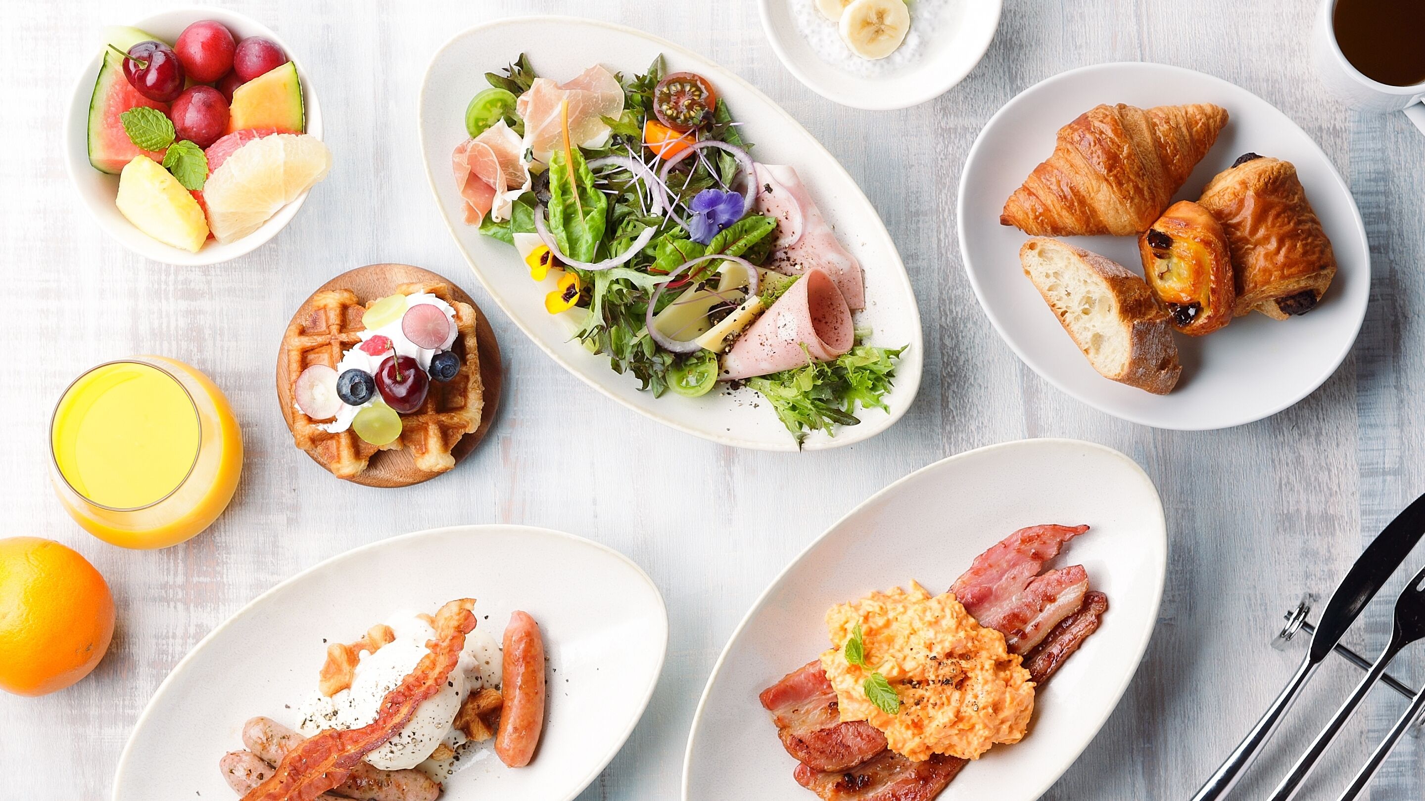 Un desayuno con diferentes platos, incluyendo un bol de fruta, ensalada, croissants, huevos revueltos, tocino y salchichas.