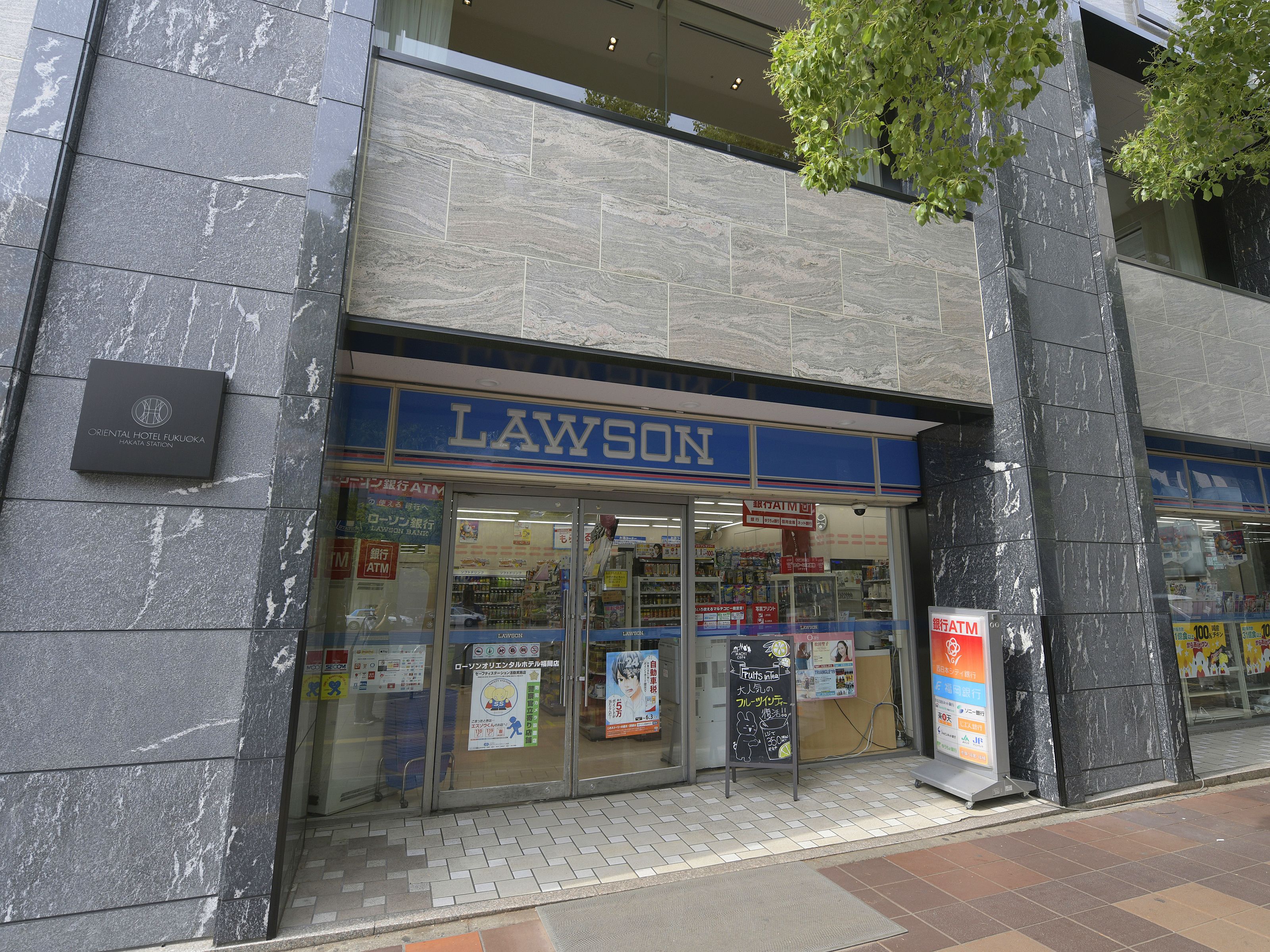 En Lawson-närbutik med en blå skylt, glasdörrar och en synlig bankomat.