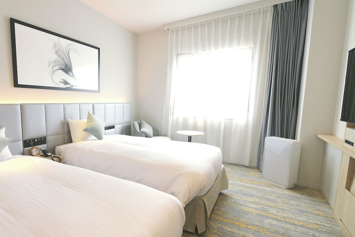 A hotel room with two beds, a bedside table, a chair, a TV, a window with curtains, and a painting on the wall.